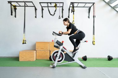 Sağlık kulübünde sabit bisiklet üzerinde egzersiz yapan kararlı genç bir kadının yan görüntüsü