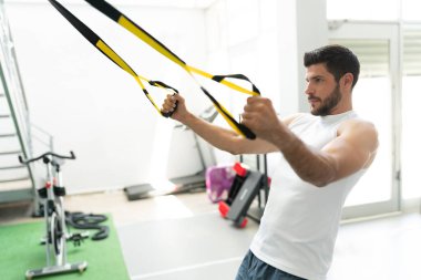 Yakışıklı Latin genç adam fitness kulübünde TRX bandosuyla egzersiz yapıyor.