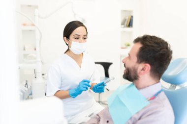 Yüz maskeli kadın doktor diş kliniğinde sandalyede oturan orta yaşlı adamı kontrol ediyor.