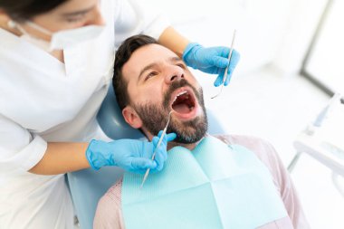 Kadın doktor, klinikte dişçi aynası ve oymacısı olan yetişkin bir adamı muayene ediyor.