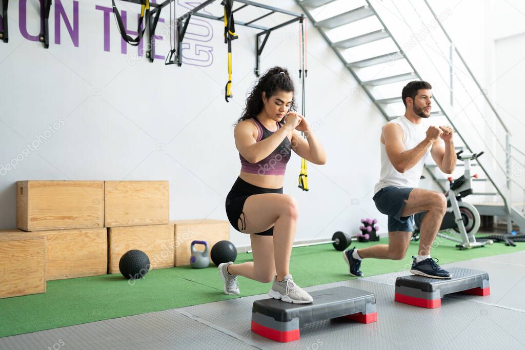 Hispanic active young man and woman doing lunges on step platforms at cross-training gym