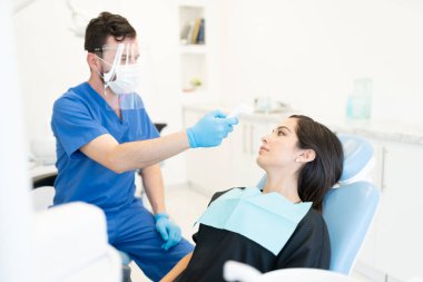 Erkek dişçi, Coronavirüs salgını sırasında odontoloji kliniğinde hastanın sıcaklığını kontrol ediyor.