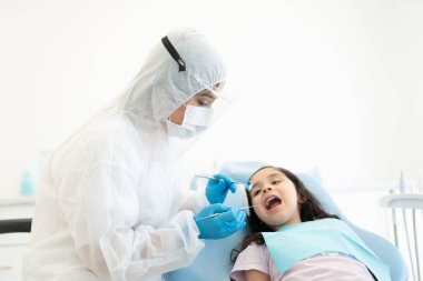 COVID19 krizi sırasında diş aynası ve oymacısı olan bir kızın dişlerini kontrol eden koruyucu iş elbisesi uzmanı.