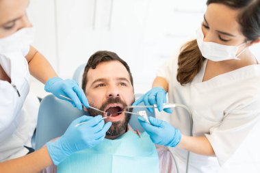 Bir çift kadın diş hekimi, dişçi kliniğinde orta yaşlı adamı aletlerle inceliyor.