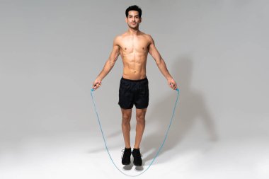 Üstsüz kaslı genç adam gri arka planda atlama halatıyla idman yapıyor.