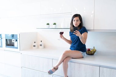 Latin genç bir kadın mutfak tezgahında otururken akıllı telefondan mesaj atıyor.