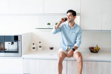 İspanyol genç adam mutfakta otururken akıllı telefondan konuşuyor.