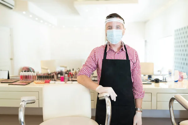 Portrait of male artist in face mask standing by chair in beauty studio during coronavirus outbreak