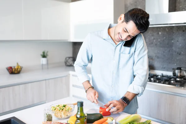 在家里切蔬菜的时候 用智能手机笑一个英俊的男人 — 图库照片