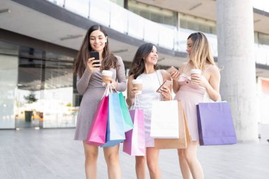 Alışveriş çantaları olan mutlu güzel kadınlar alışveriş merkezinin önünde dikilirken akıllı telefon kullanıyorlar.