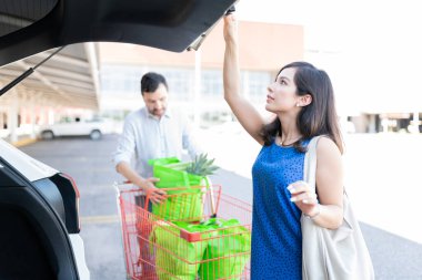 Erkek arkadaşı alışveriş arabasından poşetleri çıkarırken kadın bagajı açıyor.
