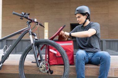 İspanyol kurye bisikletin yanında otururken sırt çantasından paketi çıkarıyor.