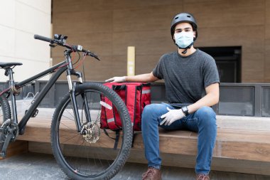 Sırt çantasında otururken maske takan genç teslimatçı.