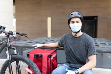 Bisikletle sırt çantasıyla otururken yüz maskesi ve kask takan teslimatçı.
