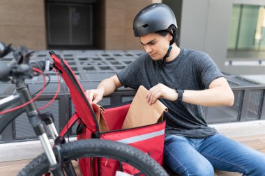 Bisikletin yanında otururken sırt çantasında paket arayan genç teslimatçı.