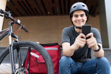 Gülümseyen İspanyol kurye bisikletle otururken mobil uygulama kullanıyor.