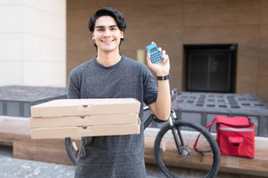 Gülümseyen yakışıklı teslimatçı elinde pizza kutuları tutarken kredi kartı okuyucusunu gösteriyor.