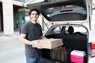 Gülümseyen yakışıklı teslimatçı araba bagajından pizza kutularını kaldırıyor.