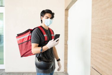 Yüz maskeli İspanyol kurye binanın kapısında dururken akıllı telefon kullanıyor.