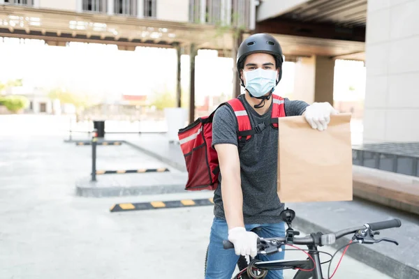 Delivery man in face mask holding parcel while sitting on bicycle in city
