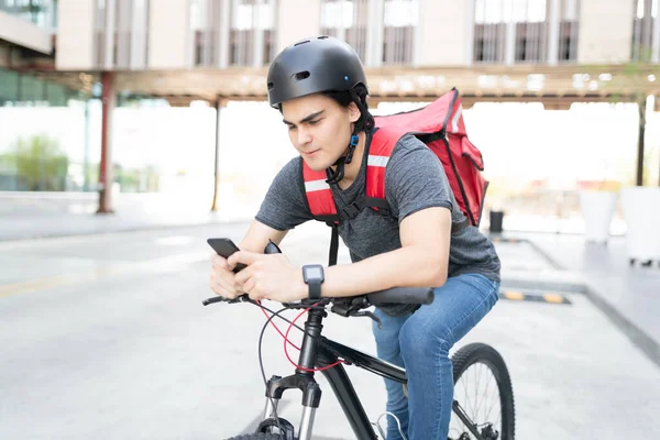 自転車に乗っている間にスマートフォンの地図を使用してハンサムな若い配達人 — ストック写真