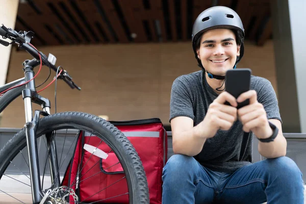 Gülümseyen İspanyol kurye bisikletle otururken mobil uygulama kullanıyor.