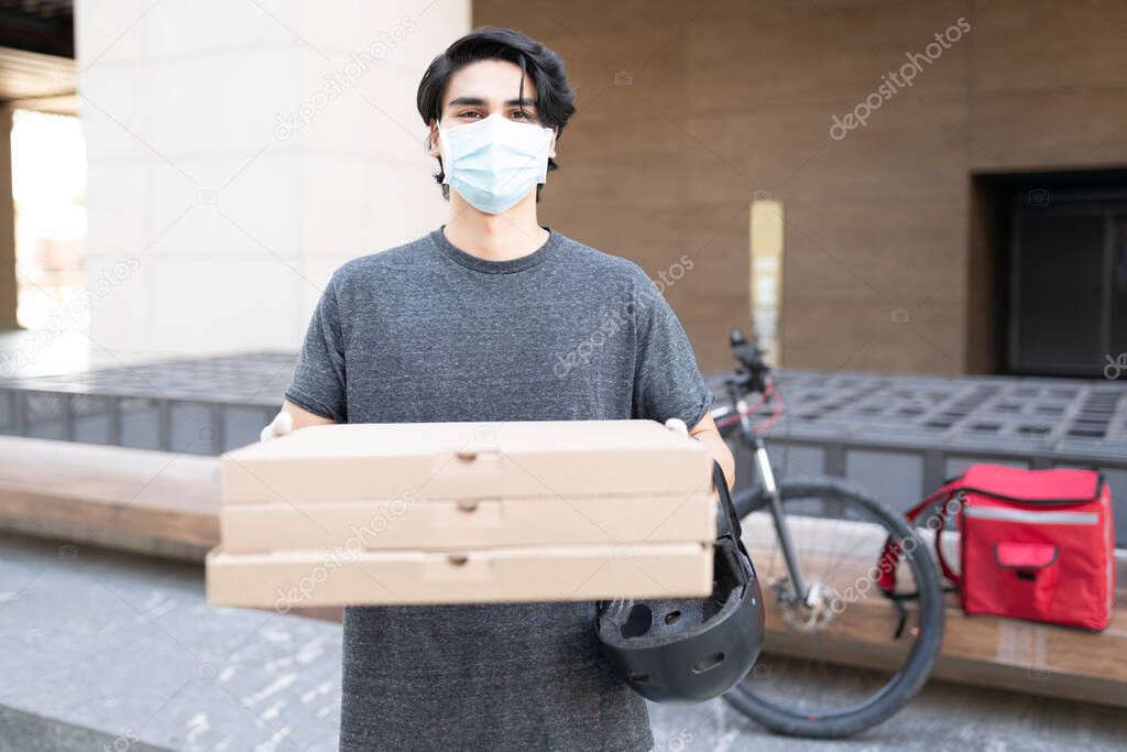 Young delivery man wearing face mask while carrying stack of pizza boxes