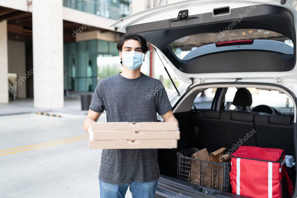 Portrait of delivery man in face mask holding pizza boxes by car trunk