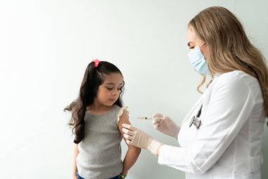 Şirin küçük kız korkmuş görünüyor ve bir doktorun ofisinde aşı olurken şırınganın iğnesine bakıyor.