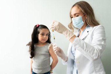 Korkmuş küçük kız şırıngaya bakarken bir doktor stüdyoda onun için aşı hazırlıyor.