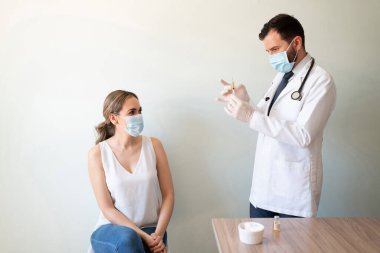 Doktorun ofisinde otururken elinde aşıyla şırınga tutan doktora bakan gergin bir kadın.