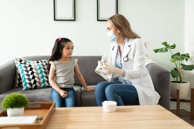 Kadın doktor küçük bir kız için aşı hazırlıyor ve daha az gergin hissetmesi için onunla konuşuyor.