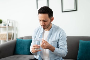 Evindeki oturma odasında otururken elinde bir bardak su ve hap tutan İspanyol bir adam.
