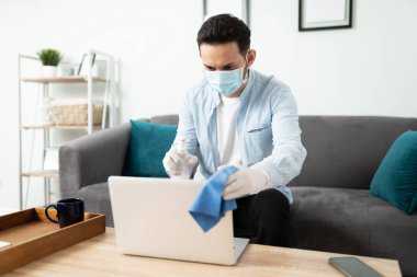Enfeksiyonu önlemek için laptopunu dezenfekte ederken yüz maskesi ve eldiven kullanan yakışıklı bir adam.