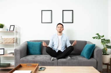 30 'lu yaşlarındaki yakışıklı İspanyol bir adam kanepede otururken huzur içinde meditasyon yapıyor.