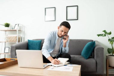 Evde bilgisayarla iş yapan yakışıklı bir adam ve meşgul görünen ve not alan bir telefon görüşmesi yapıyor.