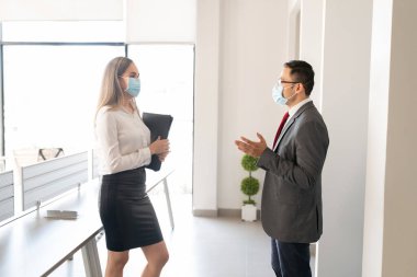 Genç iş ortakları koruyucu yüz maskesi takıyor ve sosyal mesafeyle ayakta duruyor ofiste planlarını tartışıyorlar.