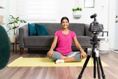 Güzel bir Latin sağlık koçunun portresi yoga minderinde oturuyor ve evden izleyicileri için bir video kaydetmeye hazır.