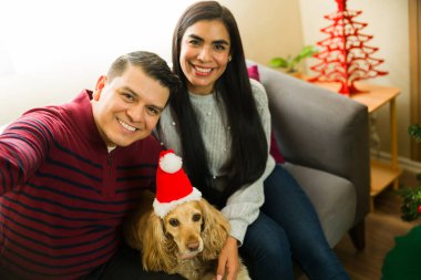 Latin çift evcil köpeğiyle selfie çekiyor. Noel şapkası takıyorlar. Tatillerde evdeki kanepede oturuyorlar.