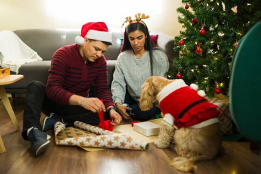 Genç, Latin bir çift Noel ağacının yanında Noel Baba kostümü giyip köpekleriyle yere Noel hediyelerini paketliyor.