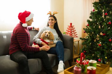Yılbaşı aksesuarları takan çiftler kış tatillerini evde geçiriyorlar, evcil köpekleriyle kanepede oturuyorlar ve şen şakrak kıyafetler giyiyorlar.