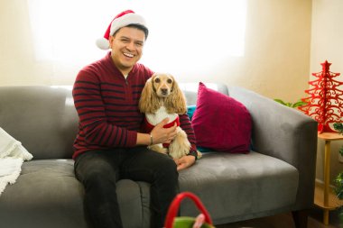 Mutlu köpek sahibi Noel Baba şapkası takıyor ve koltukta oturup Noel 'i birlikte kutlarken tüylü en iyi arkadaşına sarılıyor.