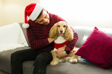 Genç İspanyol bir adam Noel şapkası takıyor. Cocker köpeğini sevgiyle kucaklıyor. Ayrıca Noel kıyafeti giyiyor.
