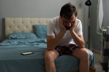 Akıllı telefonundan kötü haberler aldıktan sonra yatakta oturan üzgün adam, ayrılık sonrası depresyon ve sorunlardan muzdarip.