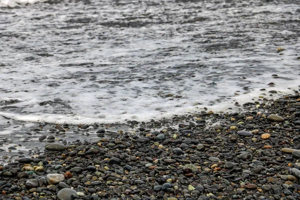 Pebbles em uma praia arenosa, pedras molhadas brilhantes e ondas. — Fotografia de Stock