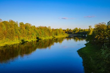 Geniş nehrin görünümü, sudaki ağaçların yansıması, doğa arka planı, gün batımı veya gün doğumu sırasında ağaçların tepesine yumuşak ışık düşüyor.