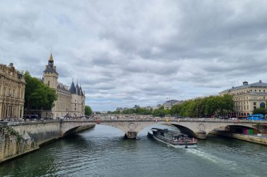Paris, Fransa, 4 Ağustos 2025: Geniş bir nehir boyunca uzanan tarihi bir köprünün manzarası, altında yavaşça geçen bir tur teknesi. İkonik, yüzyıllar öncesinin mimarisi nehir kenarlarını doldururken, verimli yeşillik doğal bir kontrastı da ekliyor. bulutlu gökyüzü