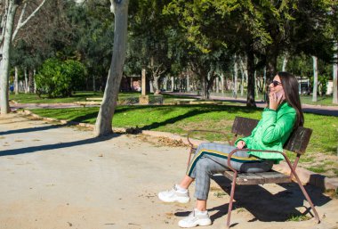 Güzel genç kadın gülümseyerek ve parkta bir bankta otururken cep telefonu kullanarak çekim.
