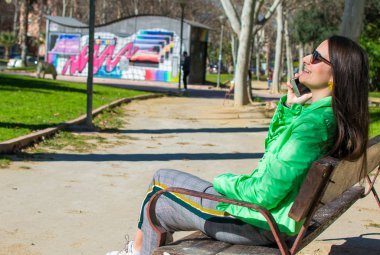 Güzel genç kadın gülümseyerek ve parkta bir bankta otururken cep telefonu kullanarak çekim.