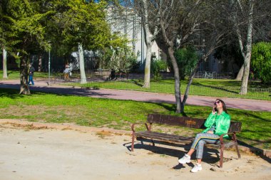 Güzel genç kadın gülümseyerek ve parkta bir bankta otururken cep telefonu kullanarak çekim.